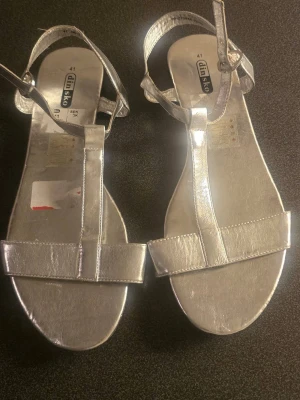 Silvriga sandaler från Din Sko - Snygga sandaler i metallic silver från Din Sko med justerbart ankelband och öppen tå. De har en stilren T-rem design och är tillverkade i ett glansigt syntetmaterial som ger en modern look. Perfekta för dig som vill sticka ut med ett par trendiga och bekväma sandaler.