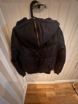 Svart pufferjacka från Marwin Sports - Svart pufferjacka från Marwin Sports med huva och dragkedja. Jackan har quiltad design och ribbade muddar nedtill. Perfekt för kalla vinterdagar och har en sportig look. Materialet är syntetiskt och jackan är vadderad för extra värme.