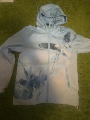 Ljusblå vindjacka med örntryck - Säljer en ljusblå vindjacka från Trapstar med stora, detaljerade örntryck på både fram- och baksidan. Jackan har huva med dragsko och dragkedja framtill. Snygg streetwear-look med tryckt Trapstar-text på ryggen. Perfekt för dig som gillar unik design.