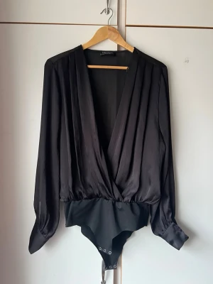 Svart body med omlott och långa ärmar - Stilren svart body från Zara Accesories med djup omlott-ringning och långa, lösa ärmar. Tillverkad i ett glansigt, mjukt tyg med knäppning i grenen. Perfekt för en trendig och elegant look.