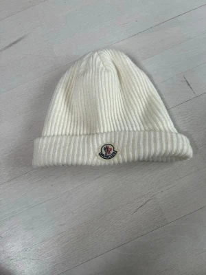 Vit ribbstickad mössa från Moncler - Säljer en stilren vit ribbstickad mössa från Moncler med uppvikt kant och klassisk logotyp framtill. Mössan är mjuk och skön, perfekt för kyliga dagar. Enkel att matcha med olika outfits och ger en clean look.