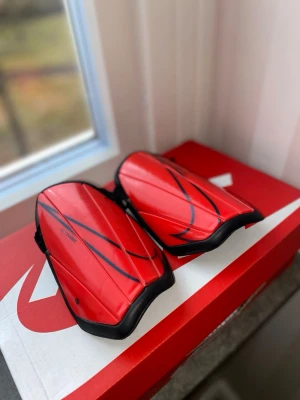 Röda benskydd från Nike - Säljer ett par röda benskydd från Nike med svarta detaljer och justerbara kardborreband. De har en sportig design och är tillverkade i plast med mjuk insida för extra komfort. Perfekta för fotbollsträning eller match.