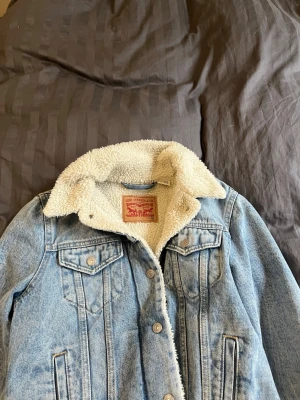 Levi's ljusblå fodrad jeansjacka - Säljer en klassisk ljusblå jeansjacka från Levi's med mysigt vitt teddyfoder. Jackan har två bröstfickor med knappar, sidofickor och silverfärgade knappar framtill. Perfekt oversized look och ikonisk Levi's-lapp i nacken. Passar dig som vill ha en tidlös streetstyle. Köppt för 1600 kr och är aldrig använd! 