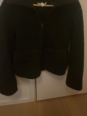 Mörk grön teddyjacka med dragkedja - Mysig mörk grön  teddyjacka med hel dragkedja framtill och hög krage. Jackan har en fluffig textur och raka ärmar, perfekt för kyliga dagar. Enkel och stilren design som passar till det mesta.