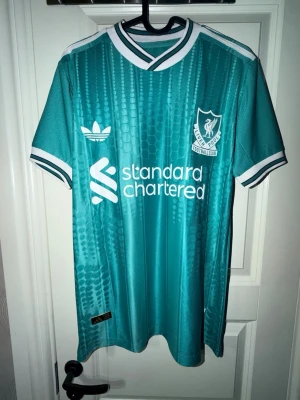 Turkos Liverpool Adidas matchtröja - Snygg turkos Liverpool FC matchtröja från Adidas med vita detaljer och klubbmärke på bröstet. Tröjan har korta ärmar, rund hals med vit och turkos ribb, samt tryckt sponsorlogga. Materialet är lätt och andas, perfekt för träning eller match.