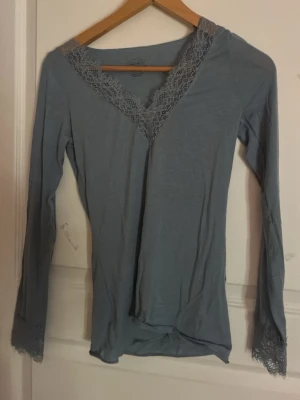 Blå långärmad loungewear med spets - Mysig blå långärmad topp med v-ringning och dekorativ spetsdetalj vid halsringning och ärmslut. Perfekt för avslappnade dagar hemma. Tunn och mjuk kvalitet som känns skön mot huden.