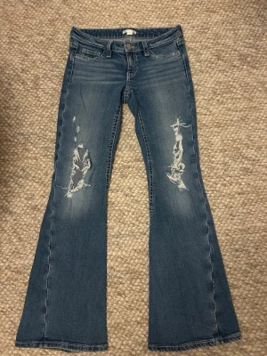 Blå bootcut jeans med hål  - Säljer ett par blå bootcut jeans med slitna detaljer på båda benen och snygga bakfickor med lock och knapp. Jeansen har låg midja och klassisk femficksdesign. Perfekta för en avslappnad och trendig look.