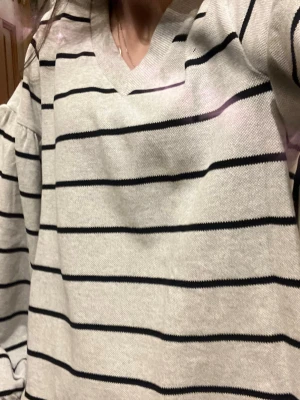 Grå randig tröja med v-ringning - Snygg grå tröja med svarta horisontella ränder och v-ringad hals. Tröjan har en avslappnad passform och långa ärmar, perfekt för en chill och stilren look. Passar till både jeans och kjol för en enkel vardagsstil. Osäker på märke, köpte i en butik hemma för 600kr. Superfin men kommer inte till användning. Knappt använd💕