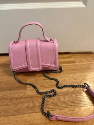 Rosa miniväska från Zara med kedja - Söt rosa axelremsväska från Zara i croco-mönstrat konstläder. Väsk manageable storlek med både handtag och kedja för axeln. Perfekt för att lyfta en outfit med en poppig färg och trendig design. Stängs med lock och har svart innerfoder.