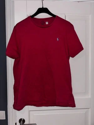 Rosa-röd t-shirt från Polo Ralph Lauren - Klassisk rosa-röd t-shirt från Polo Ralph Lauren med den ikoniska lilla loggan broderad på bröstet. Rund halsringning och korta ärmar. Perfekt basic-plagg för alla tillfällen och enkel att matcha med jeans eller shorts.