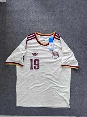 Spanien Lamine Yamal matchtröja - Vit Spanien fotbollströja från Adidas med vinröda och gula detaljer, nummer 19 och Lamine Yamal på ryggen. Tröjan har officiella märken, broderad klubbmärke och Adidas-logga, samt tryck på ärmarna. Tillverkad i lätt och ventilerande material.