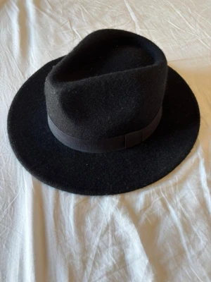 Svart filthatt med brett brätte - Stilren svart filthatt med klassiskt brett brätte och dekorativt band runt kronan. Perfekt accessoar för att lyfta din outfit med en tidlös touch. Hatten har en markerad form och är tillverkad i mjukt filtmaterial.