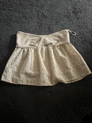 Vit broderad skort med resårmidja - Supersöt vit skort med broderade blommor och hålmönster. Skön resår i midjan med struktur och liten knytning. Perfekt för en somrig och fräsch look. Materialet känns lätt och luftigt, och modellen är både bekväm och trendig.