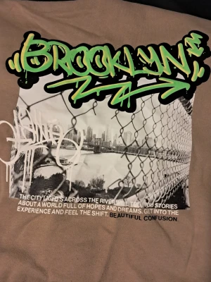 Beige hoodie med Brooklyn-tryck - Säljer en beige hoodie med dragkedja och huva. På bröstet finns ett vitt graffiti-inspirerat tryck och på ryggen ett stort Brooklyn-motiv med grön text och svartvit stadsbild. Hoodien är i mjuk bomullsmix och har fickor framtill.