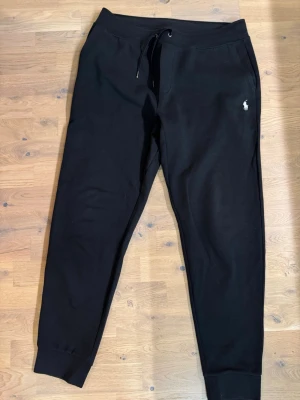 Svarta mjukisbyxor från Polo Ralph Lauren - Svarta mjukisbyxor från Polo Ralph Lauren med dragsko i midjan och ribbade muddar vid bensluten. Klassisk vit logga broderad på vänster höft. Sköna och stilrena byxor i mjukt material, perfekta för en avslappnad look.
