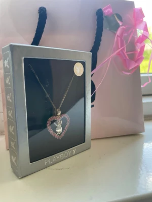 Playboy halsband med hjärthänge - Säljer ett snygg vintage halsband från Playboy med ett silverfärgat hjärthänge dekorerat med rosa stenar och den klassiska kaninloggan i mitten. Kedjan är tunn. Kommer i originalförpackning och med en rosa presentpåse. Har mörka fläckar men aldrig använts