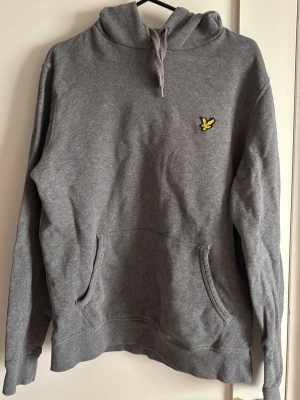 Grå hoodie från Lyle & Scott - Säljer en grå hoodie från Lyle & Scott med klassisk gul broderad örnlogga på bröstet. Hoodien har huva med dragsko och en stor magficka. Tillverkad i mjukt bomullsmaterial och har ribbade muddar vid ärmar och nederkant. Perfekt för en avslappnad stil.