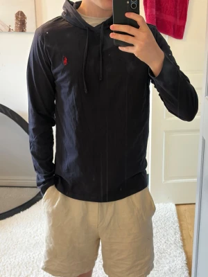 Mörkblå hoodie från Polo Ralph Lauren - Snygg mörkblå hoodie från Polo Ralph Lauren med röd broderad logga på bröstet. Hoodien har dragsko i huvan och är tillverkad i ett mjukt, tunt material som passar perfekt för en avslappnad stil. Långärmad och med klassisk passform.