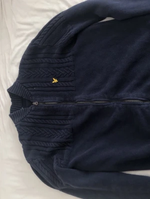 Mörkblå stickad ziptröja Lyle & Scott - Mörkblå stickad ziptröja från Lyle & Scott med snygga flätstickade detaljer upptill och ribbad krage. Tröjan har dragkedja framtill och den klassiska gula loggan på bröstet. Perfekt för lager-på-lager och stilren look.