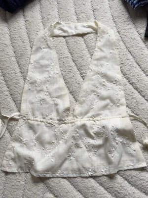 Vit broderad omlottopp - Söt vit omlottopp med broderade blommor och knytband i midjan. Toppen är ärmlös och har en djup v-ringning, perfekt för varma dagar. Lätt och luftig känsla med fina detaljer.