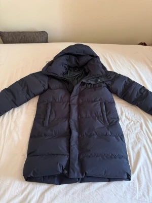 Everest Parka - Snygg svart parka från Everest med stor huva och två djupa fickor framtill. Jackan har en quiltad design med breda paneler och stängs med dragkedja och tryckknappar. Perfekt för kalla vinterdagar och ger en riktigt varm känsla.