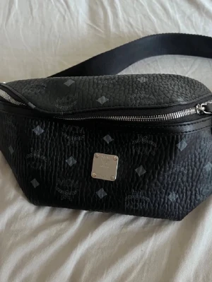 Svart magväska från MCM - Snygg svart magväska från MCM i präglat läder med silverfärgad dragkedja och logotypdetaljer över hela väskan. Justerbart band för perfekt passform. Perfekt accessoar för att hålla koll på dina grejer med stil.
