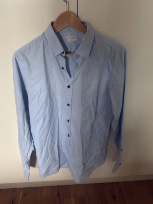 Ljusblå skjorta från Selected Homme - Snygg ljusblå skjorta från Selected Homme i slim fit-modell. Skjortan har klassisk krage, svarta knappar och långa ärmar. Perfekt för dig som gillar stilrena och enkla plagg med modern passform.