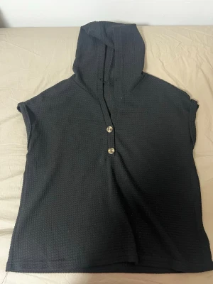 Svart kortärmad hoodie med knappar - Svart hoodie i ett strukturerat tyg med huva och två knappar framtill. Modellen är kortärmad och har en avslappnad passform, perfekt för en chill och stilren look.