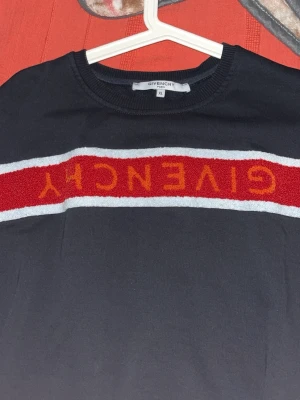 Svart Givenchy t-shirt med röd logga - Svart t-shirt från Givenchy med en bred röd och vit rand över bröstet där Givenchy-loggan står i rött. Klassisk rund halsringning och korta ärmar. Materialet känns mjukt och bekvämt, perfekt för en trendig streetwear-look. Storlek s och m finns 