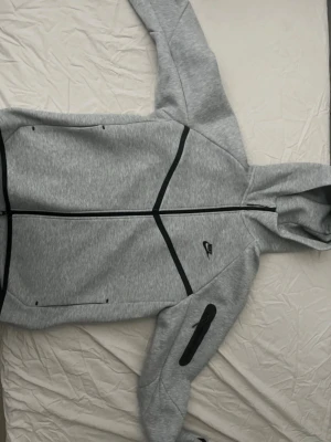 Grå Nike Tech Fleece hoodie - Snygg grå Nike Tech Fleece hoodie med svarta detaljer och dragkedja framtill. Hoodien har huva, två fickor och en extra ficka med dragkedja på ärmen. Perfekt för en sportig och stilren look. Använd en ända gång