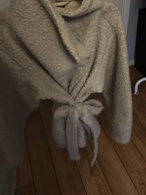 Beige teddyjacka med knytband - Supermjuk och fluffig teddyjacka i beige med bred krage och knytband i midjan. Jackan har långa ärmar och en oversized känsla, perfekt för att hålla sig varm och snygg. Materialet är teddy/fuskpäls som ger en mysig look.