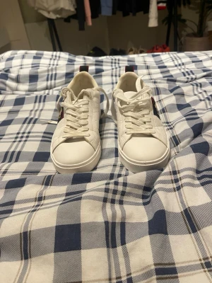 Vita sneakers från pier one  med brodyr - Snygga vita sneakers från pier one med klassisk låg profil. Skorna har broderad patch i rött, blått och guld på sidan samt präglat mönster i lädret. Tjock vit sula och vita skosnören. Perfekta för dig som vill sticka ut med exklusiva detaljer.