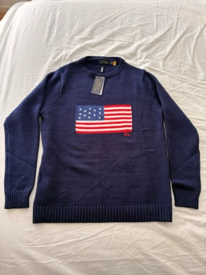 Mörkblå stickad tröja Polo Ralph Lauren - Mörkblå stickad tröja från Polo Ralph Lauren med amerikansk flagga framtill. Tröjan har rund halsringning och ribbstickade muddar. Perfekt för dig som gillar klassisk och stilren design.