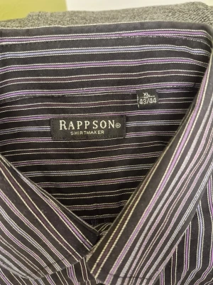 VINTAGE randig skjorta från Rappson XL - Snygg vintage skjorta från Rappson i svart med tunna vita och lila ränder. Klassisk krage, långärmad och knäppning framtill. Perfekt för dig som gillar stilrena och moderna skjortor med lite edge.