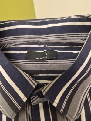 Randig skjorta från Selected, XL - Snygg skjorta från Selected i storlek XL med blå, vita och grå vertikala ränder. Skjortan har klassisk krage, långa ärmar och knäppning framtill. Perfekt för en stilren look och passar till många olika outfits.