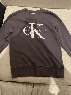Svart sweatshirt från Calvin Klein Jeans - Snygg svart sweatshirt från Calvin Klein Jeans med stort vitt CK-tryck på bröstet. Tröjan har rund halsringning, långa ärmar och ribbade muddar vid ärmslut och nederkant. Perfekt för en stilren och avslappnad look.
