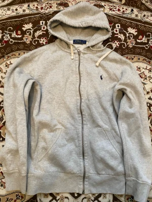 Grå hoodie från Polo Ralph Lauren - Snygg grå hoodie från Polo Ralph Lauren med dragkedja, huva och kängurufickor. Klassisk broderad logga på bröstet och vita snören i huvan. Mjuk bomullsblandning som är perfekt för en avslappnad stil.