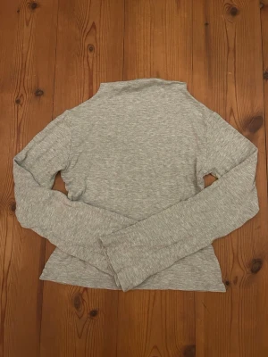 Grå polotopp från Lager 157 - Snygg och enkel grå polotopp från The Black Label Collection. Toppen är långärmad och har en tight passform med mjukt material. Perfekt att styla med jeans eller kjol för en clean look.