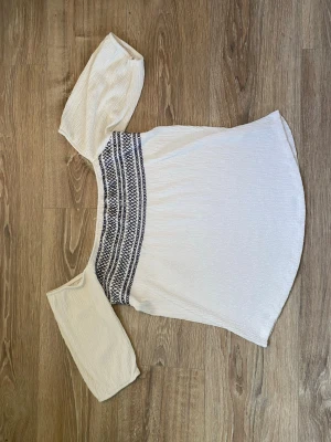 Vit offshoulder blus med brodyr - Trendig vit offshoulder blus med svarta broderade detaljer över bröstet. Blusen har vida, korta ärmar och är gjord i ett lätt, luftigt tyg som ger en avslappnad och somrig känsla. Perfekt att styla med jeans eller kjol för en cool look.