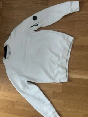 Vit sweatshirt från C.P. Company - Snygg vit sweatshirt från C.P. Company med rund hals och lång ärm. Tröjan har en svart logodetalj på ena ärmen som ger en cool touch. Tillverkad i mjukt bomullsmaterial för skön komfort. Perfekt för en stilren och avslappnad look.