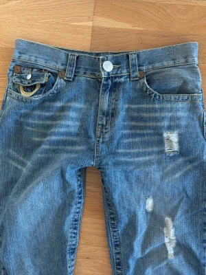 True Religion slitna blå jeans - Snygga blå jeans från True Religion med slitningar framtill och på bakfickorna. Perfekta för en avslappnad streetstil. Säljer pg av att dom är för korta för mig som är 175cm, hon jag köpte dom av har sytt upp dom. Dom är cr 100cm i längd  