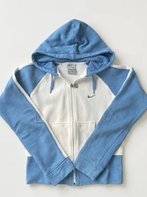 Nike Air hoodie i blått och vitt - Nike Air hoodie i mjuk bomullsblandning med dragkedja framtill. Färgblockad design i ljusblått och vitt, broderad Nike-logga på bröstet och stor Nike Air-brodyr på ryggen. Huva med dragsko och känguruficka. Perfekt för en sportig och avslappnad stil. Passar XS eller 15 år (170) 