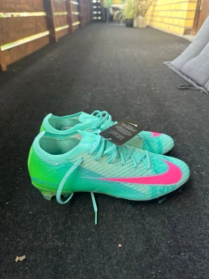 Nike Mercurial turkosa fotbollsskor - Snygga Nike Mercurial fotbollsskor i turkos med ett stort rosa Nike märke på ovansidan. Skorna har en strumpliknande passform och snörning för bra stöd. Tillverkade i lätt syntetmaterial för optimal bollkontroll och grepp på planen. Verkligen en dröm sko. Storlek 39 men känns mindre ! Pris kan diskuteras och bud! De är helt nya med prislapp. Man får med 4 bandage bindor , 1 par beskydd, 1 fotbollspåse, 2 par grepp strumpor och 1 liten Nike väska. 
