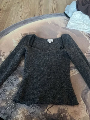 Mörkgrå stickad topp från H&M - Mörkgrå stickad topp från H&M med lång ärm och snygg, något hjärtformad urringning. Tröjan har en figurnära passform och ribbstickad struktur som ger en trendig look. Perfekt för lager-på-lager-stil under kyliga dagar.