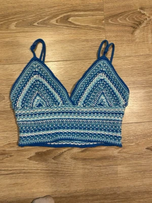 Blå virkad croppad linnetopp - Säljer en handvirkad croppad linnetopp i olika blå nyanser med smala axelband. Toppen har ett randigt mönster och trekantig form framtill. Perfekt för sommaren och ger en chill boho-vibe.