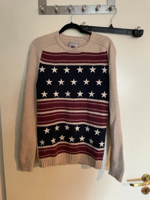 Stickad tröja med stjärnor Lexington - Stickad tröja från Lexington med beige ärmar och rygg, samt framstycke i marinblått med vita stjärnor och vinröda ränder. Klassisk rund halsringning och ribbstickade muddar. Perfekt för dig som gillar amerikansk preppy-stil.
