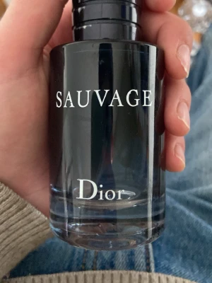 Tom dior flaska - Säljer en elegant parfymflaska från Dior, modellen Sauvage. Flaskan är cylindrisk och har en mörkblå till svart ton med vit text. Lock i matchande färg och stilren design som känns lyxig och modern 