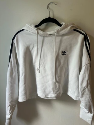 Kort vit hoodie från Adidas  - Snygg vit hoodie från Adidas med svarta ränder längs ärmarna och broderad logga på bröstet. Hoodien har huva med dragsko och är tillverkad i mjukt material. Perfekt för en sportig och avslappnad stil.