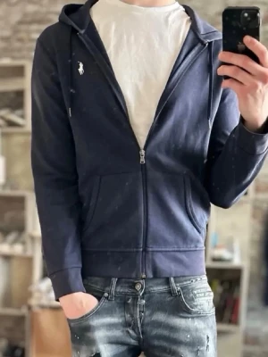 Marinblå hoodie från Polo Ralph Lauren - Snygg mörkblå hoodie med dragkedja från Polo Ralph Lauren. Klassisk design med vit logga på bröstet, huva med dragsko och två fickor framtill. Tillverkad i mjukt bomullsmaterial som är skönt att bära. Perfekt till jeans eller joggers.