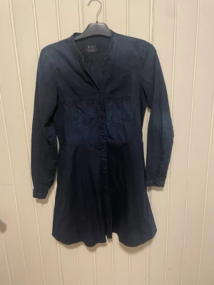 Mörkblå skjortklänning från edc - Säljer en snygg mörkblå skjortklänning från edc med lång ärm och knappar hela vägen framtill. Klänningen har en enkel, rak krage och är tillverkad i ett mjukt bomullsmaterial. Perfekt för dig som gillar en avslappnad men ändå stilren look.